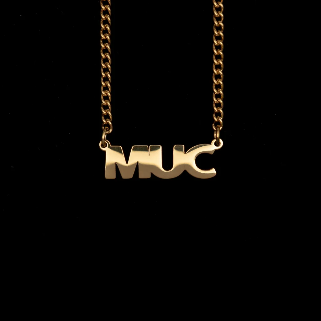 Goldfarbene Kette mit dem Schriftzug-Anhänger „MUC“ vor schwarzem Hintergrund