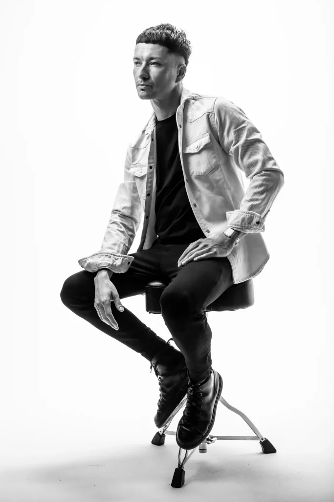 Genji Yoshida in heller Jacke auf einem Hocker sitzend, Schwarzweiß-Aufnahme im Studio