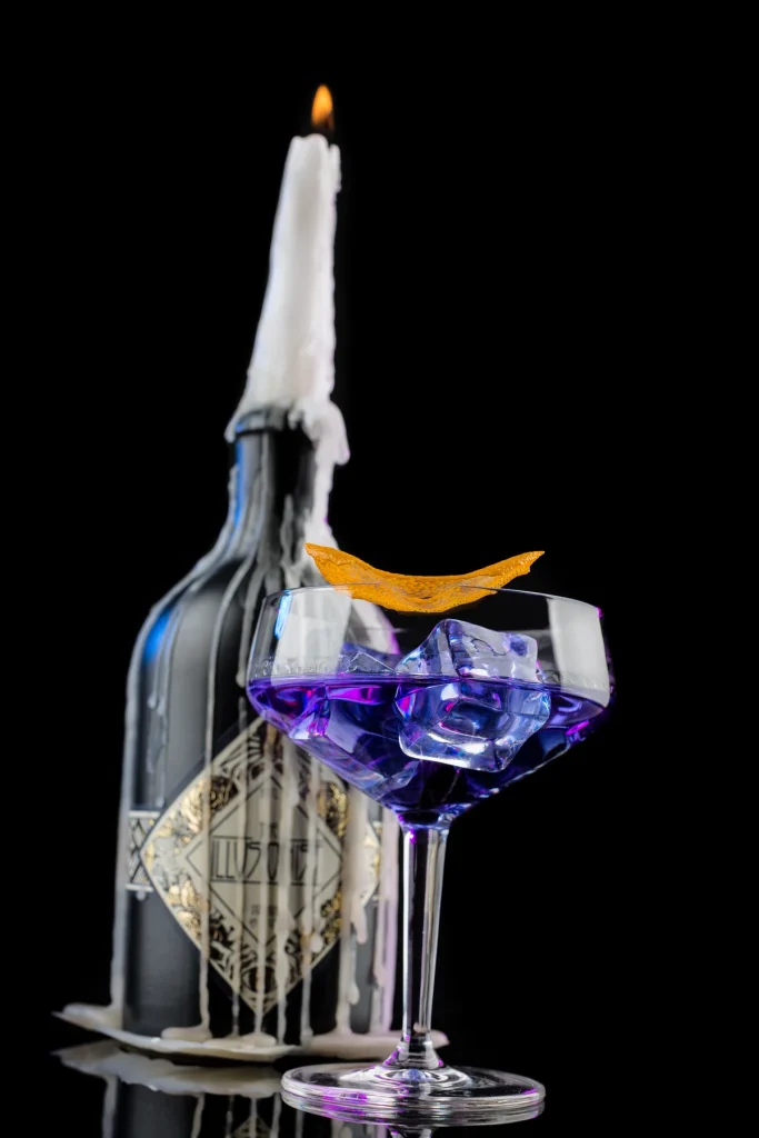 Cocktail im Coupeglas mit Eiswürfeln vor einer Flasche mit brennender Kerze und herabgelaufenem Wachs