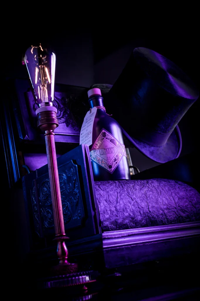 Flasche am Polstersessel, daneben eine Glühbirne auf Ständer und ein Zylinderhut in violettem Licht