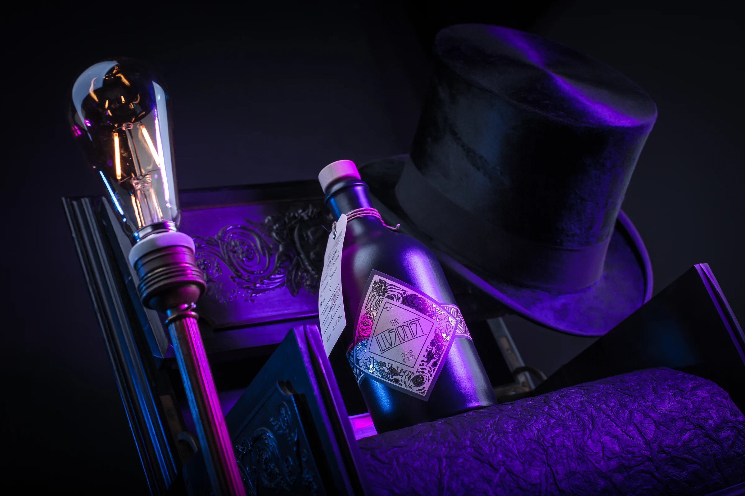 Flasche „The Illusionist Dry Gin“ zwischen Zylinder, Glühbirne und Polstermöbel, violett beleuchtet
