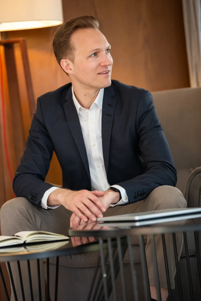 Johannes Rauch im Sakko blickt aus dem Bild, im Vordergrund ein Tisch mit Notizbuch und Laptop