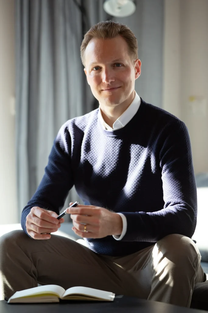Johannes Rauch sitzt mit Stift in der Hand auf einem Sofa, vor ihm ein Notizbuch, Blick in die Kamera